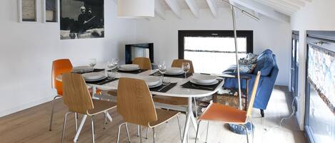 Un comedor con una mesa blanca dispuesta para cuatro personas, rodeada de sillas de madera. Un televisor está montado en la pared, y hay una ventana que proporciona luz natural.