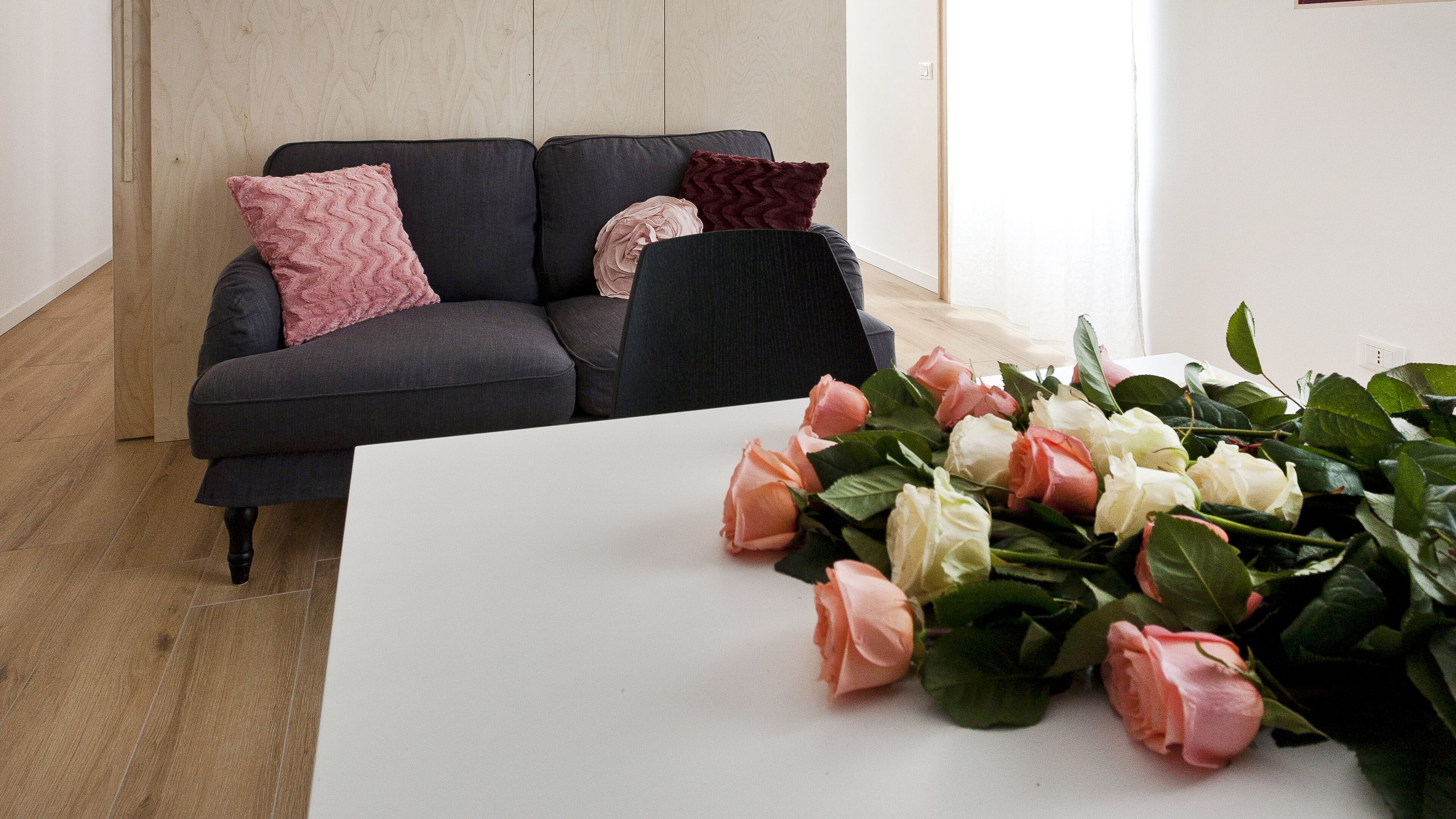 Una sala de estar moderna con un sofá gris oscuro, dos almohadones rosados y uno borgoña, y una mesa blanca con un ramo de rosas.