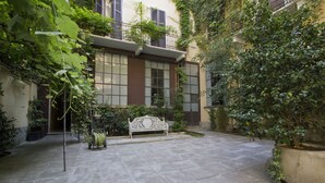 A courtyard with a bench, potted plants, and a building with large windows and balconies. - Italianway   - Caretto (Milan)