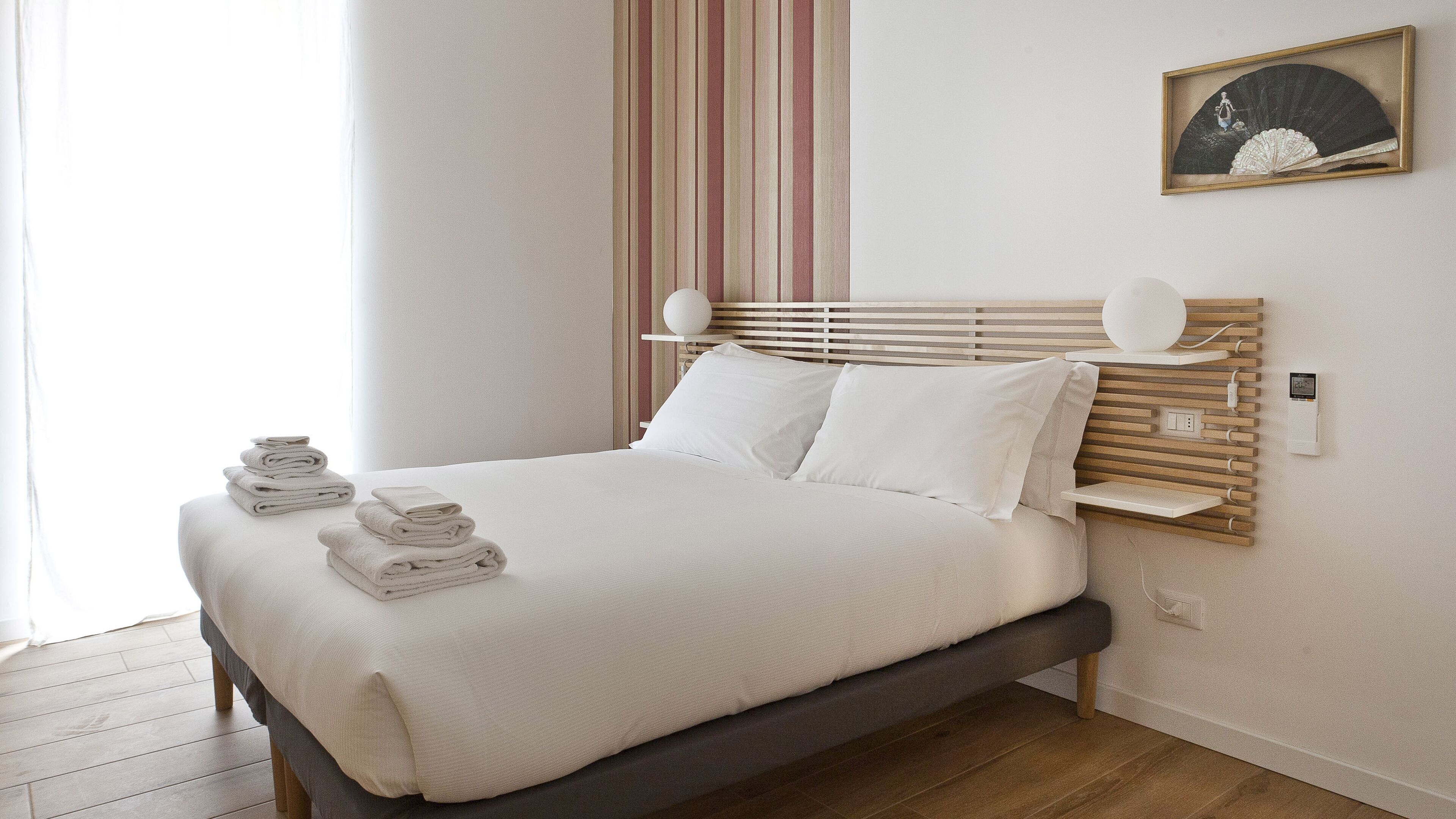 Un lit bien fait, avec du linge de lit blanc et des oreillers, une tête de lit en bois, une table de chevet avec une lampe et une prise de courant fixée au mur.