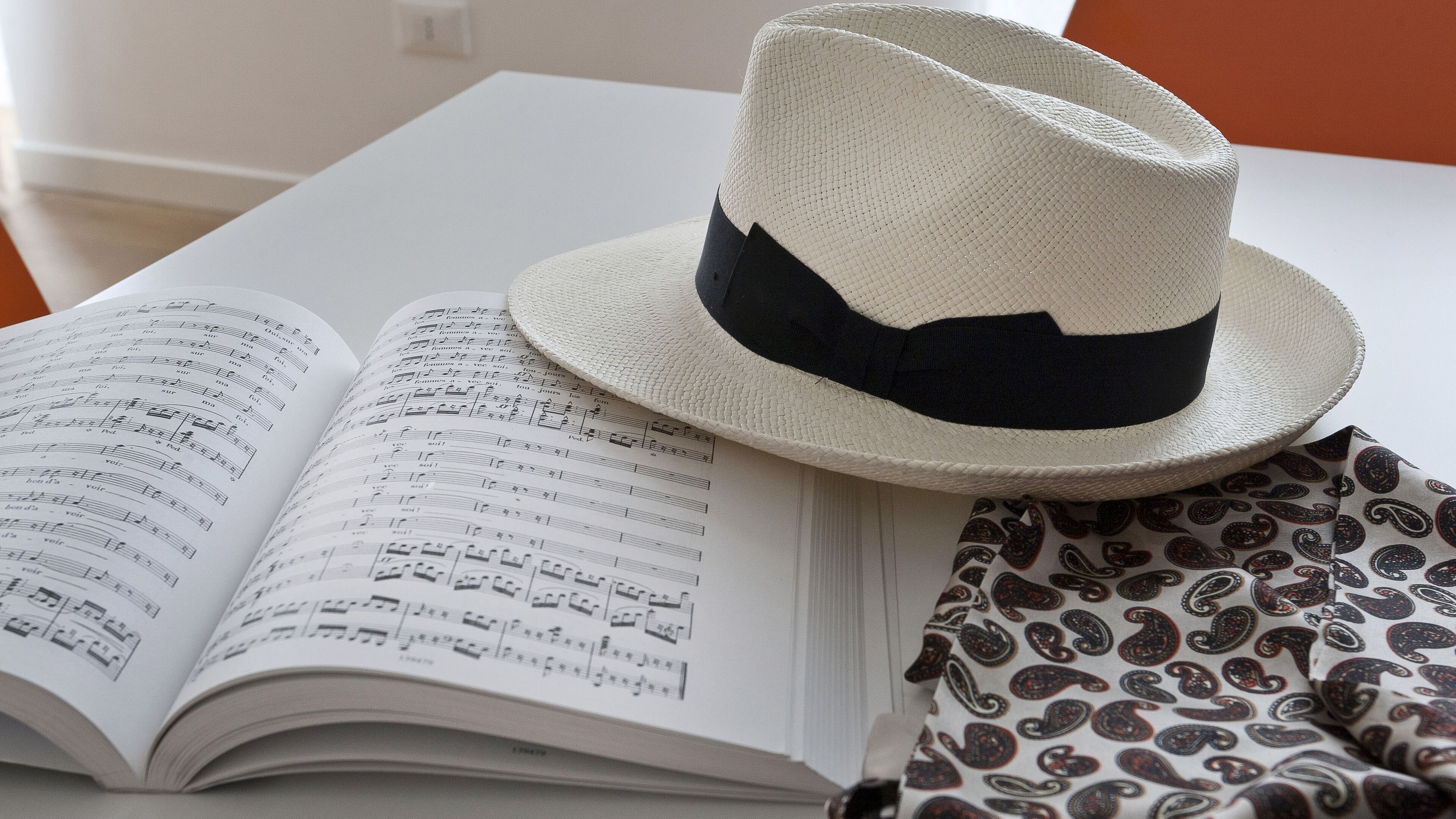 Un libro abierto con partituras sobre una mesa, un sombrero y un tejido estampado.