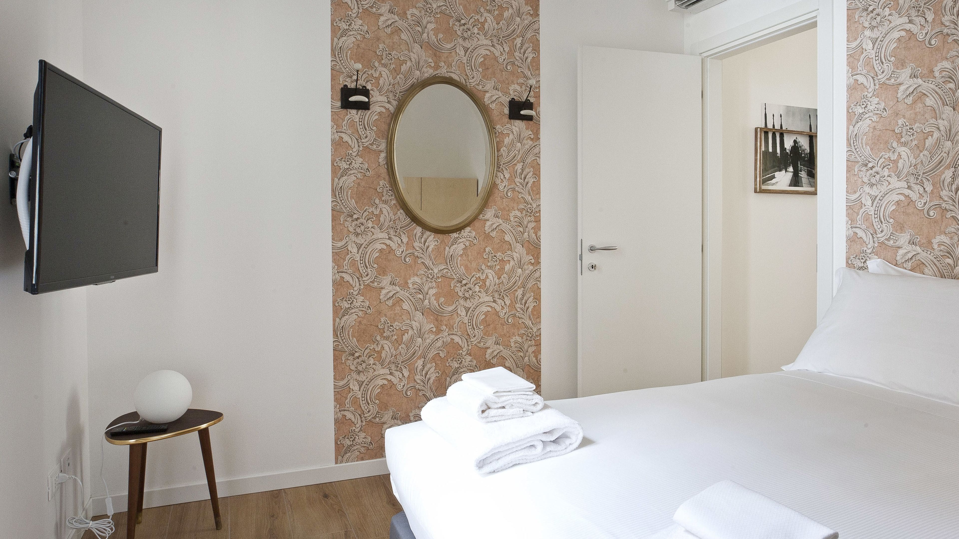 A hotel room with a bed, a bedside table with a lamp, a mirror, and a wall-mounted TV.