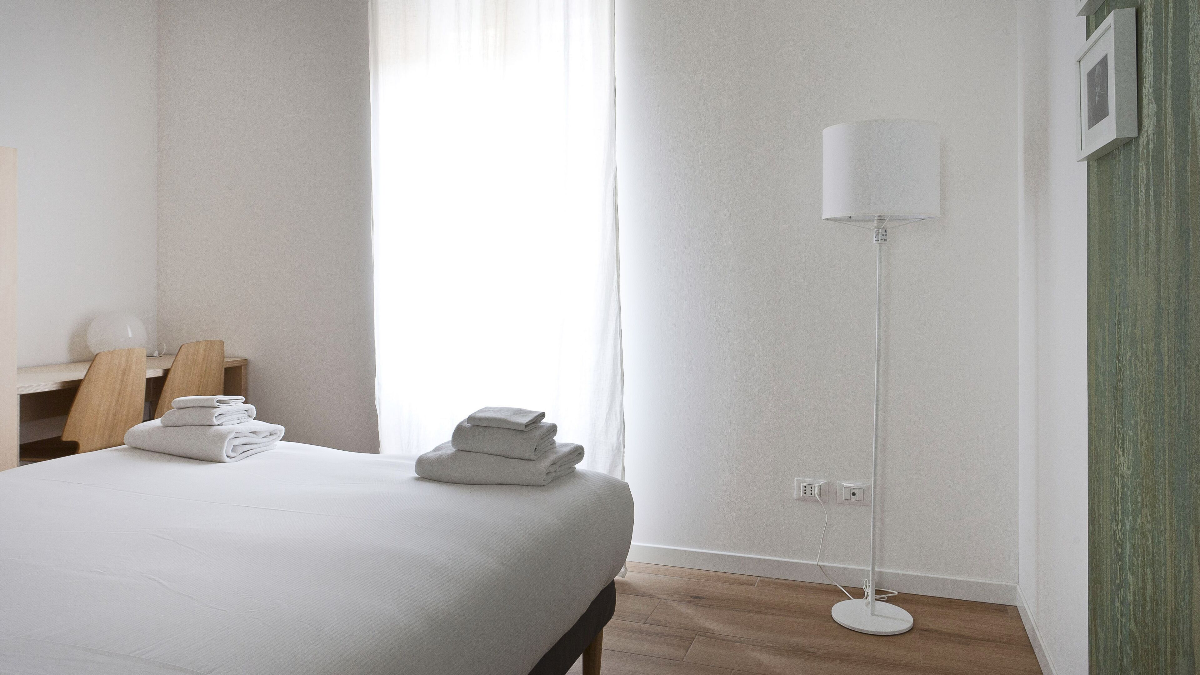 A hotel room with a neatly made bed, a bedside table with a lamp, a chair, and a floor lamp.