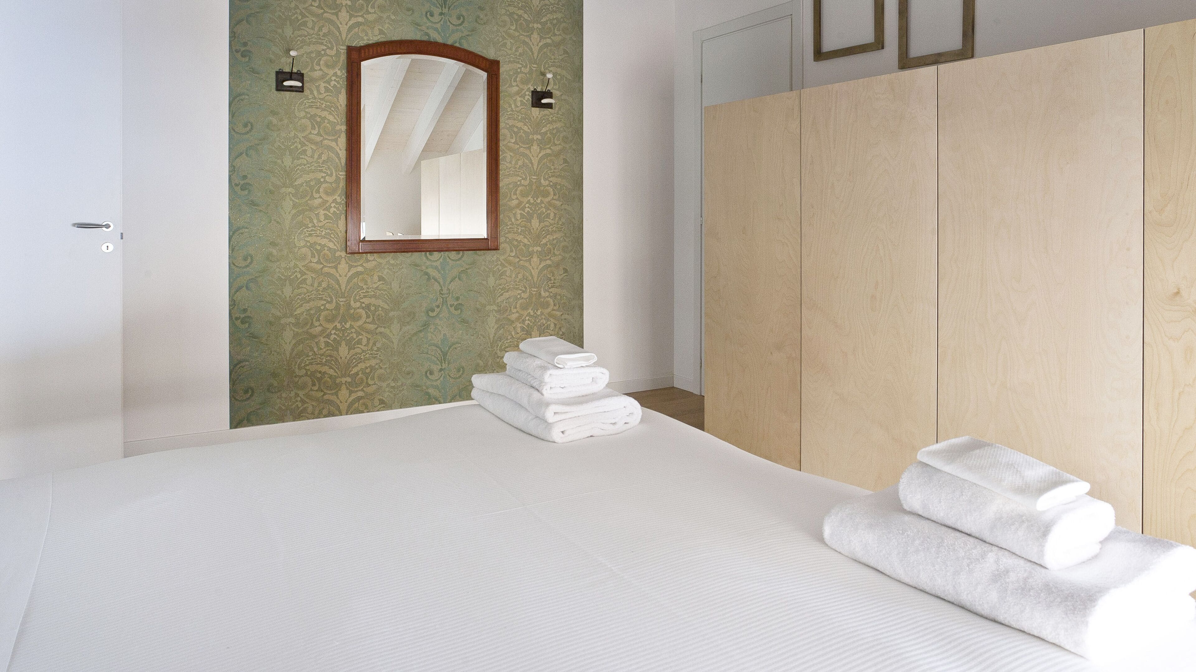Una habitación de hotel con una cama, un espejo, un armario de madera y una pila de toallas blancas.