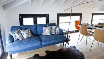 A modern living room with a blue sofa, patterned cushions, a dining table with chairs, and a large window with a view.