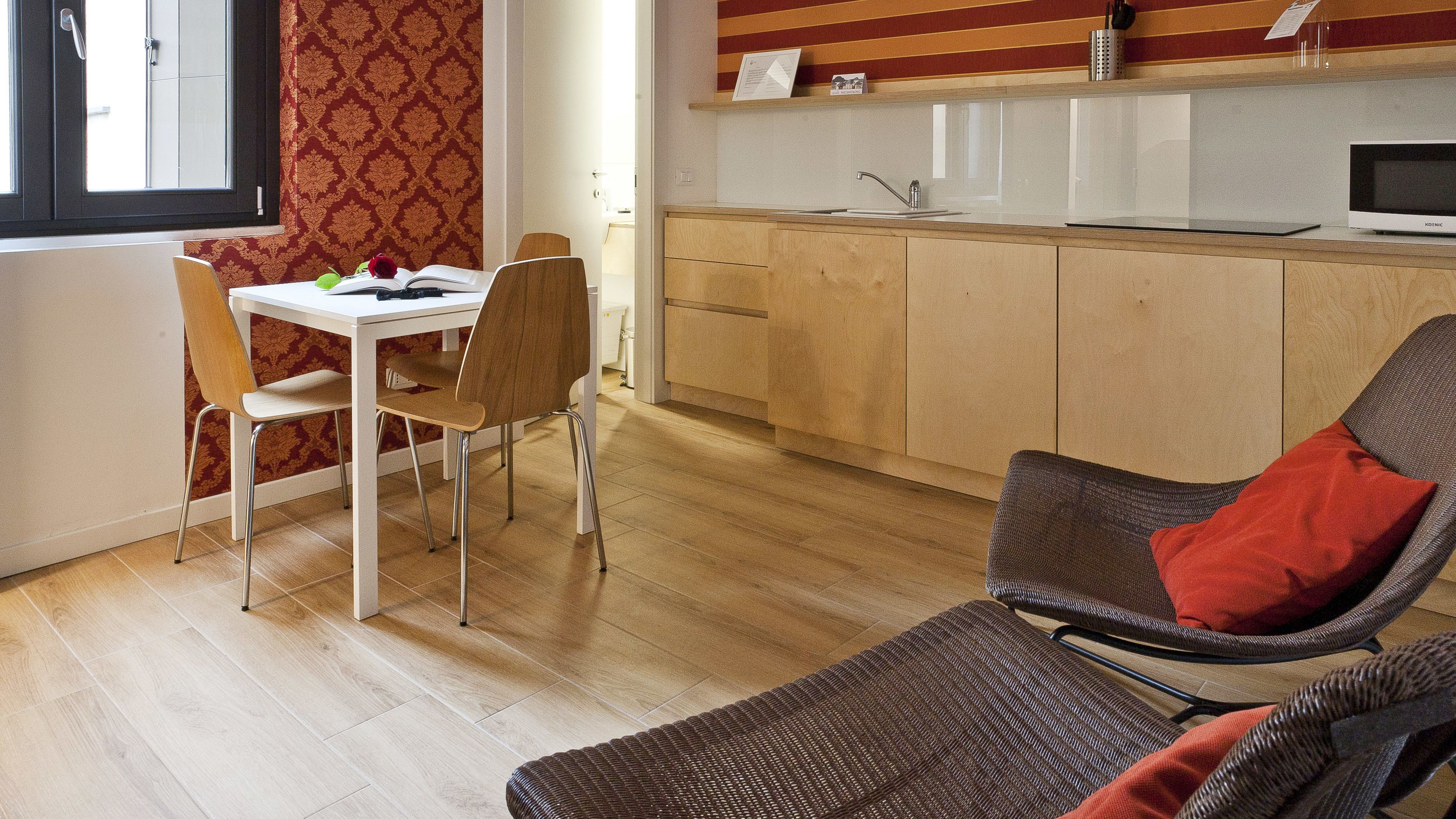 Una cocina moderna con armarios de madera, encimera blanca y un microondas. Hay una zona de comedor con una mesa y sillas, y una silla de mimbre con un cojín rojo.