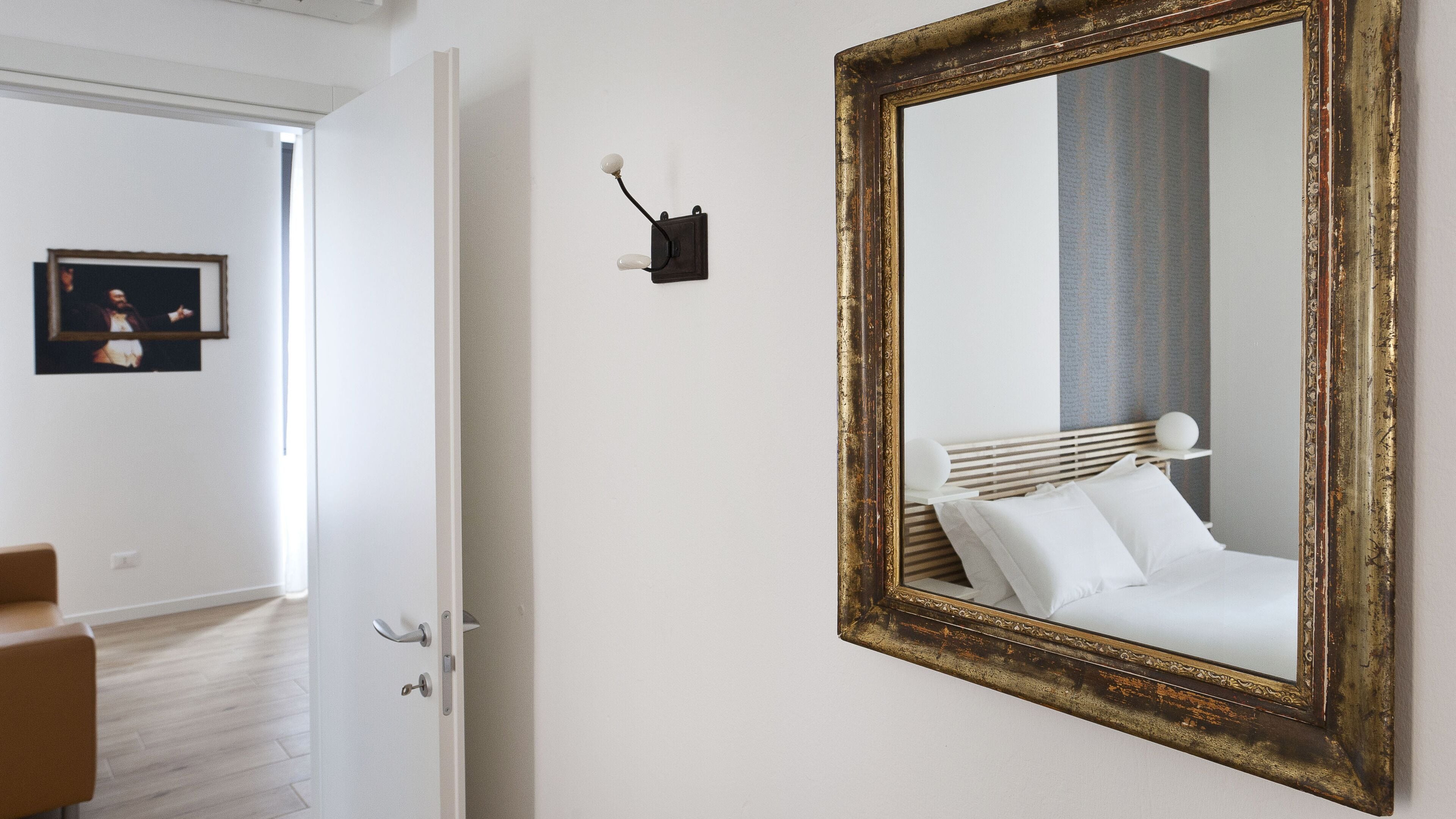 Habitación de hotel con un espejo grande, una cama con ropa de cama blanca, una mesita de noche con lámpara y un cuadro en la pared.