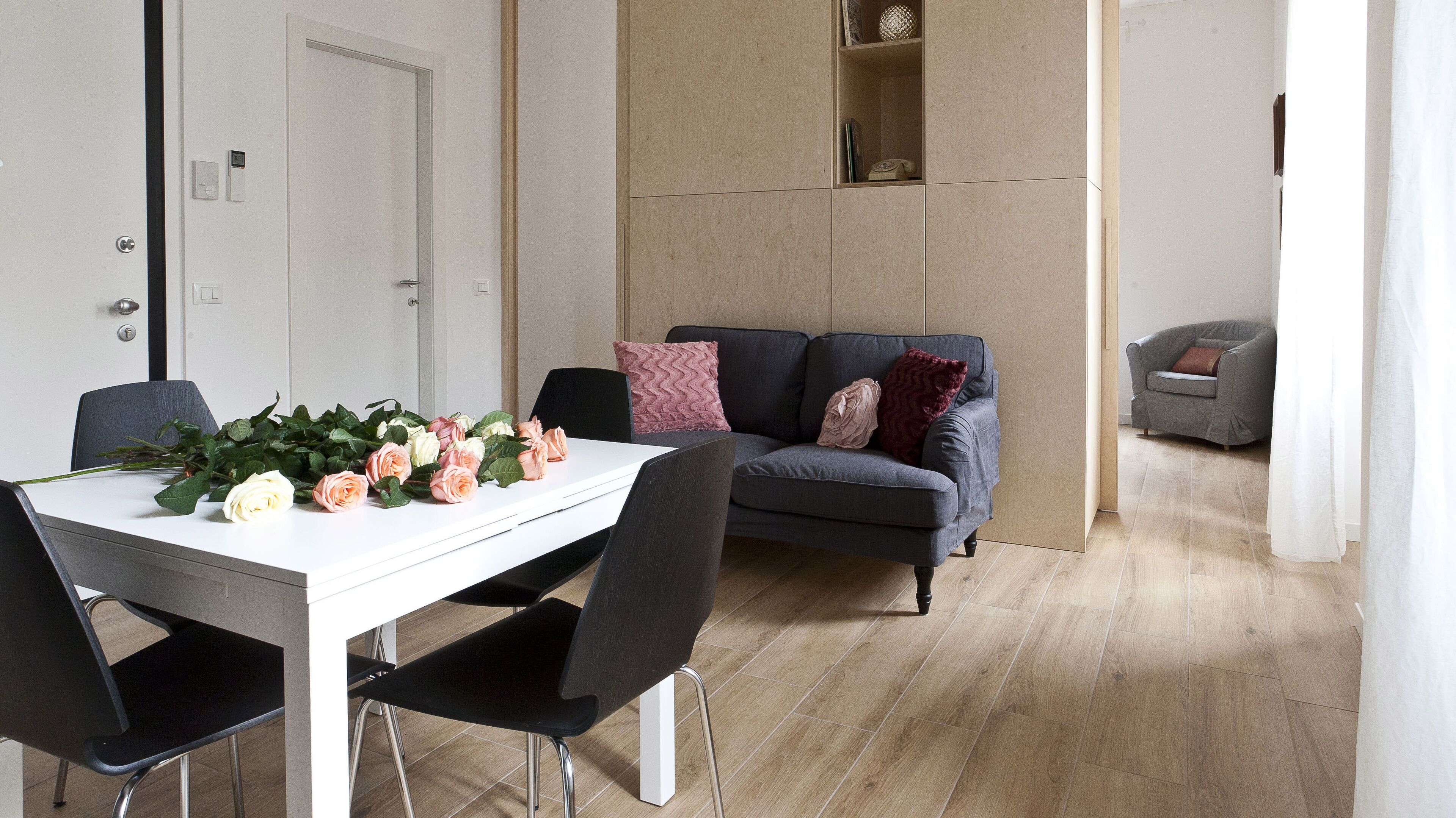 A modern living room with a white dining table, black chairs, a grey sofa with patterned cushions, and a wooden floor.