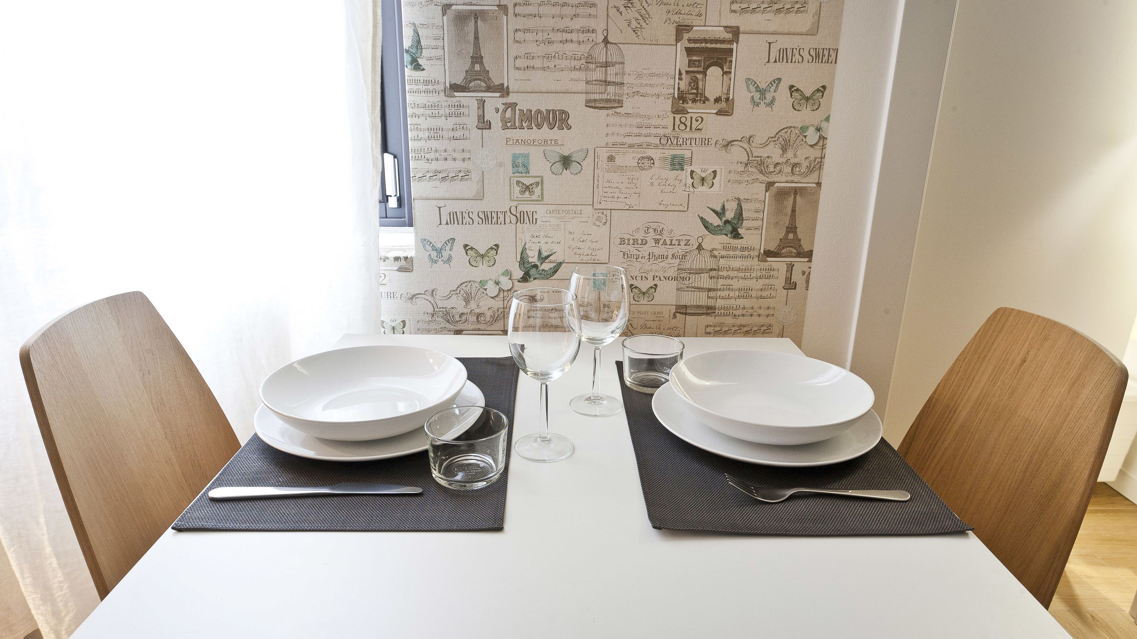 A dining table set with two white plates, two wine glasses, and cutlery, against a wall with vintage wallpaper.
