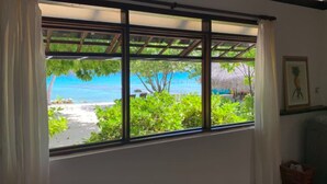 Una habitación con un ventanal grande con vista a la playa, un cuadro en la pared y una cesta tejida en el suelo.