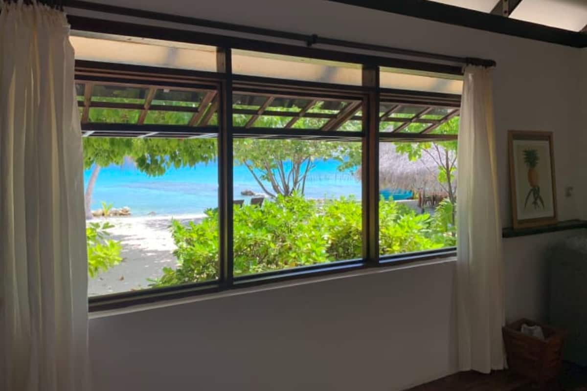 Quarto com uma grande janela com vista para a praia, um quadro na parede e uma cesta de palha no chão.