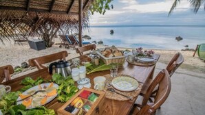 Mesa de comedor dispuesta con platos, cubiertos y una cesta de frutas, con vista a una playa que cuenta con una estructura de techo de paja y sillones de descanso.