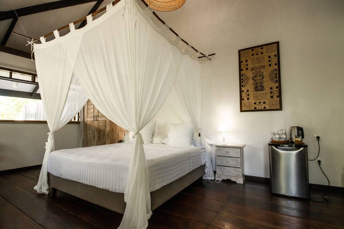 Quarto com cama de dossel, mesa de cabeceira com abajur, armário pequeno e lixeira.