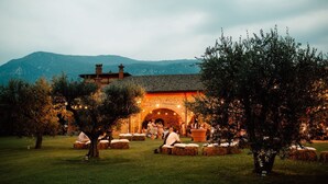 Un espacio de comedor al aire libre con fardos de heno y árboles, un edificio de piedra con entrada arqueada y un paisaje montañoso de fondo.