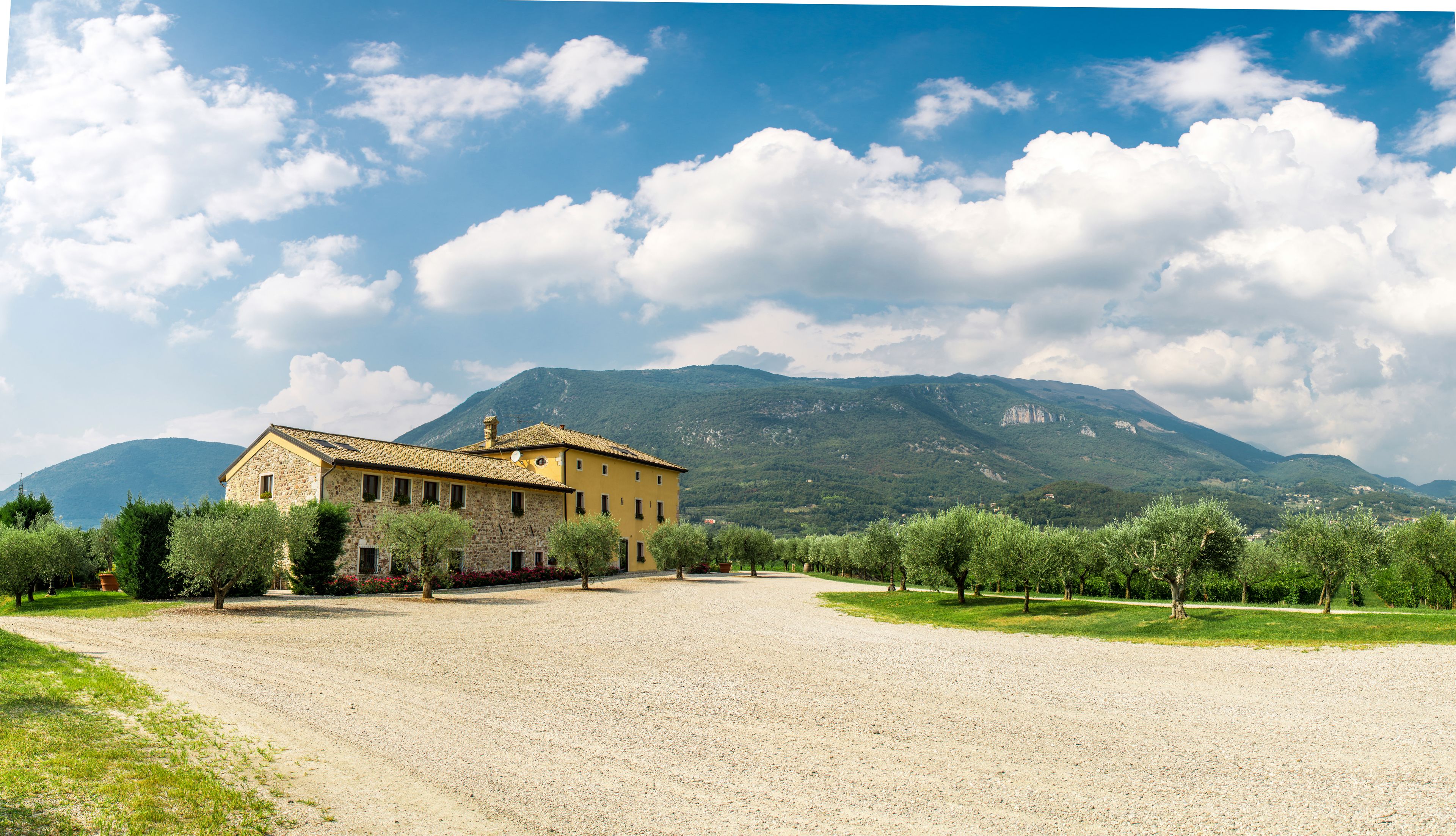 A rustic stone building with a gravel driveway, surrounded by olive trees and a mountainous backdrop.