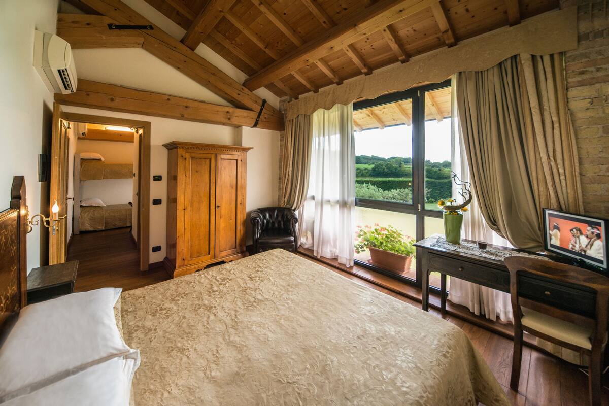 Een slaapkamer met een groot bed, houten balken in het plafond, een bureau met stoel, een televisie en een raam met gordijnen.