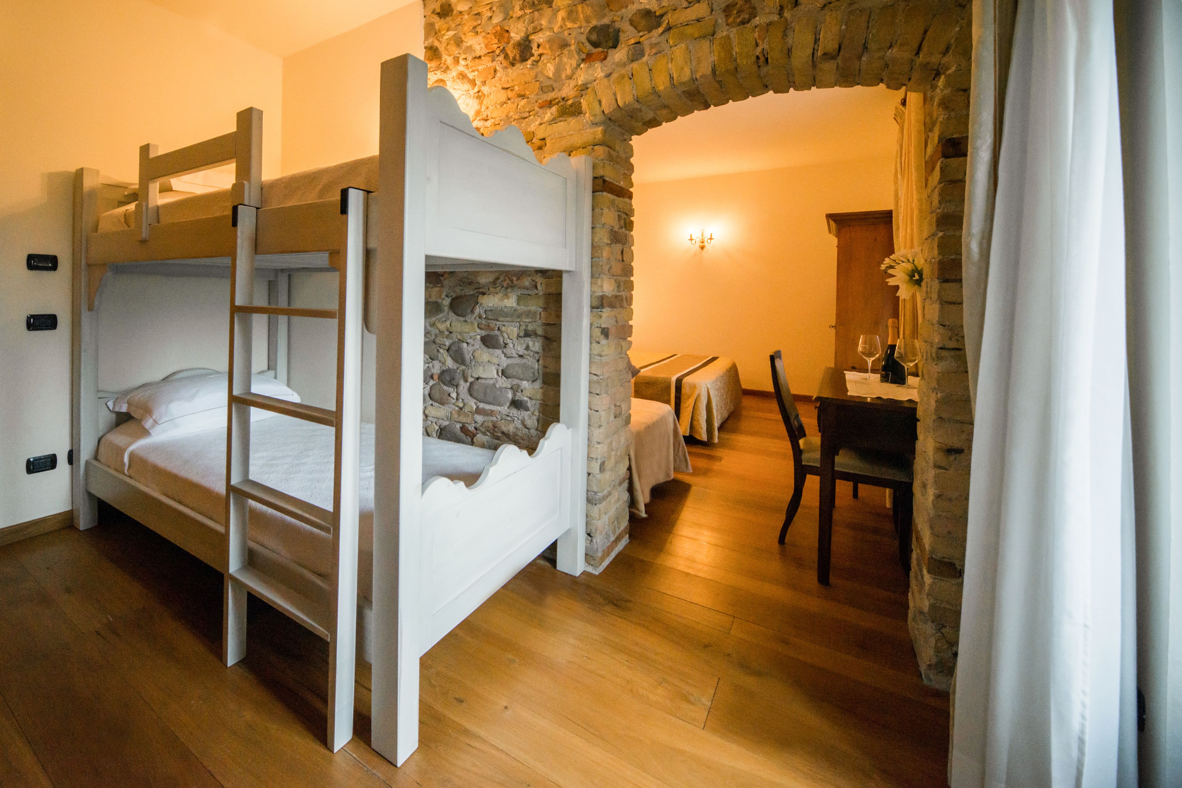 A bunk bed with a ladder, a stone wall, a wooden desk, and a chair in a room with wooden flooring.