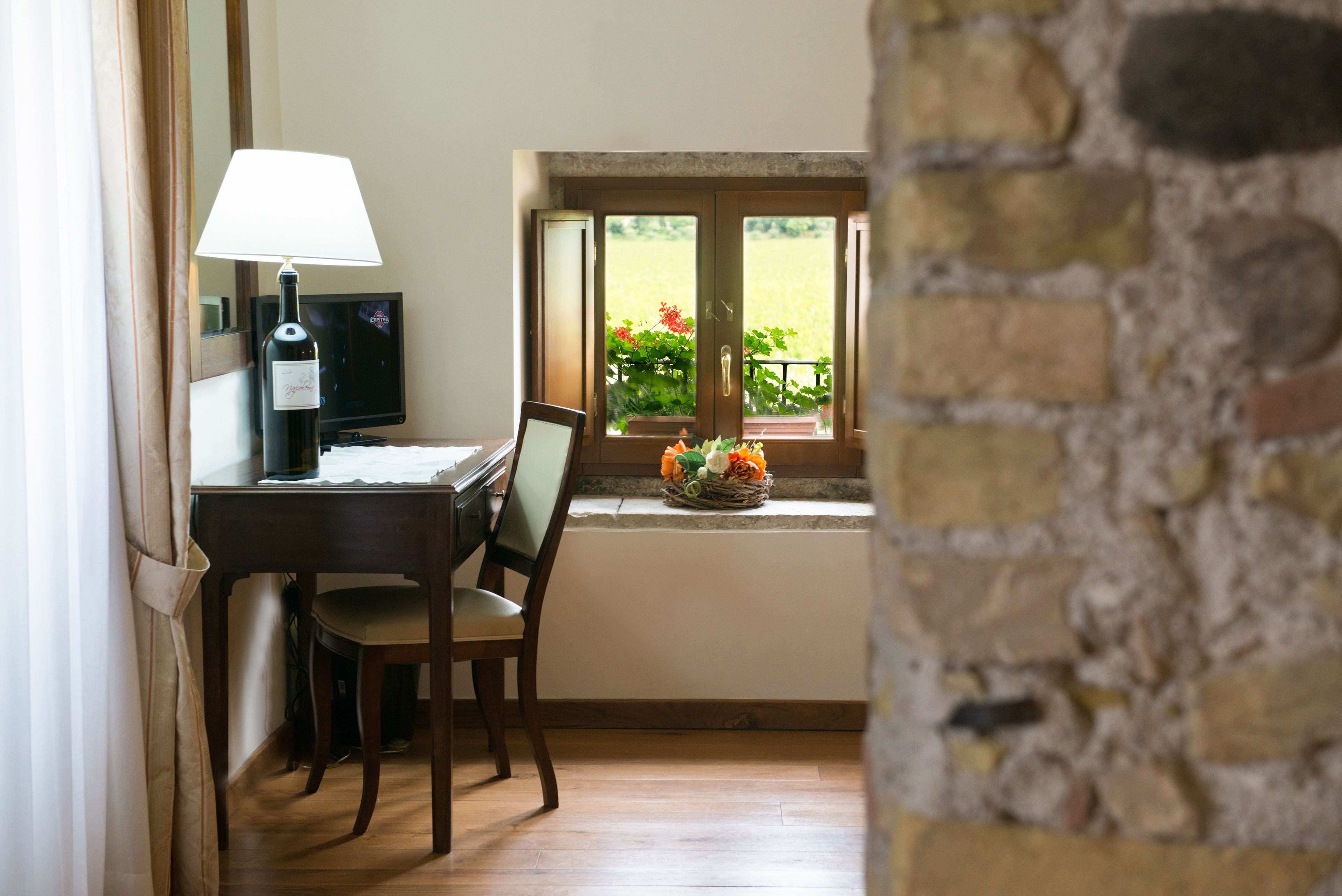 Ein Zimmer mit Schreibtisch, Stuhl, Lampe, Fernseher und einem Fenster mit Blick ins Grüne.