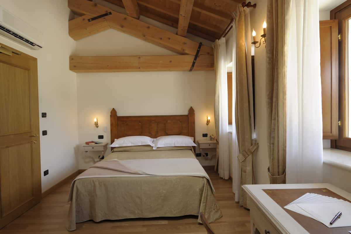 Een slaapkamer met een houten plafond, een bed met een beige dekbedovertrek, nachtkastjes met lampen, en een raam met gordijnen.