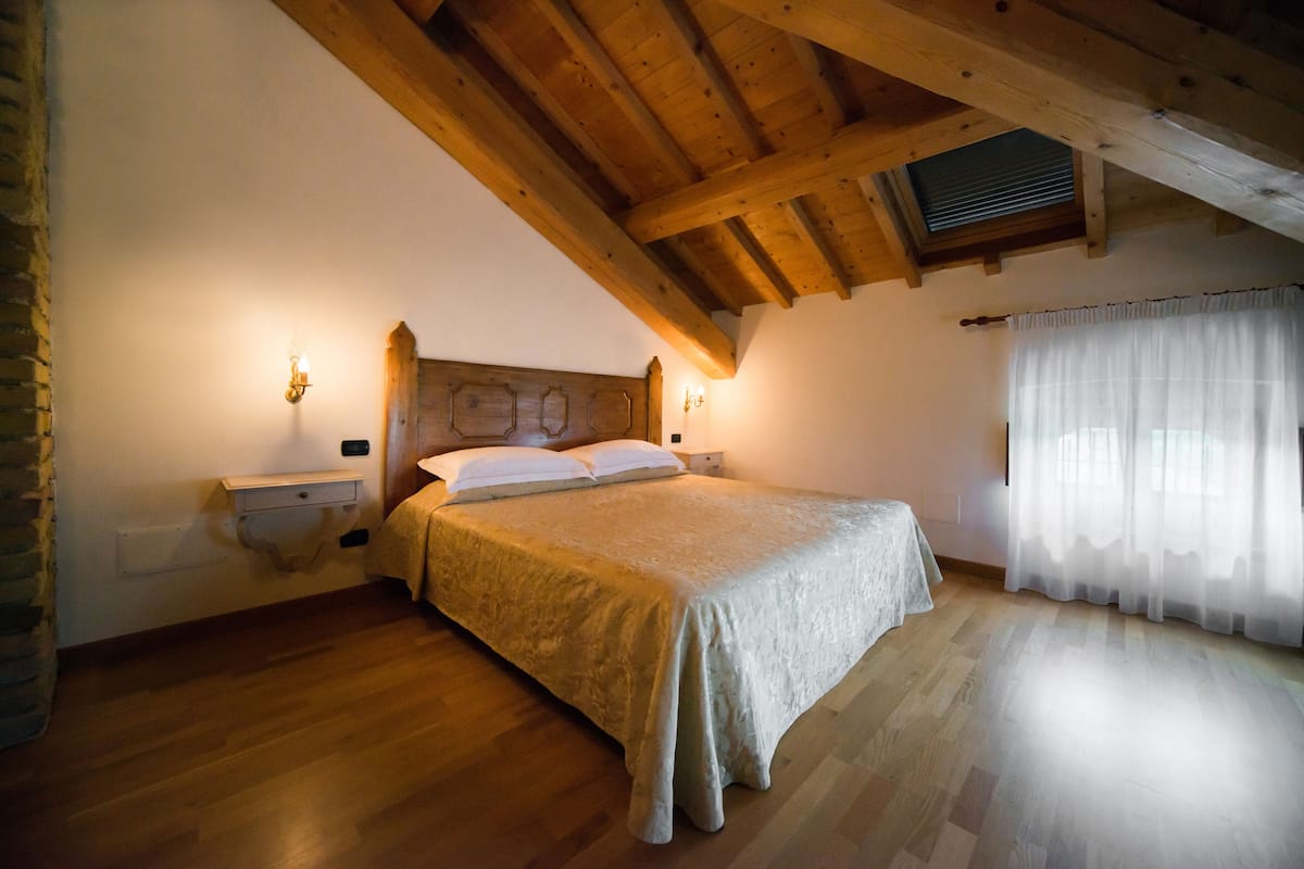 Een slaapkamer met een houten plafond, een bed met een witte dekbedovertrek, twee nachtkastjes en een raam met dunne gordijnen.