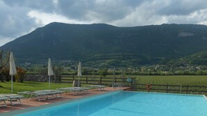 Piscina con sillas de descanso y sombrillas, rodeada por un muro de piedra y vistas a una montaña y un pueblo.