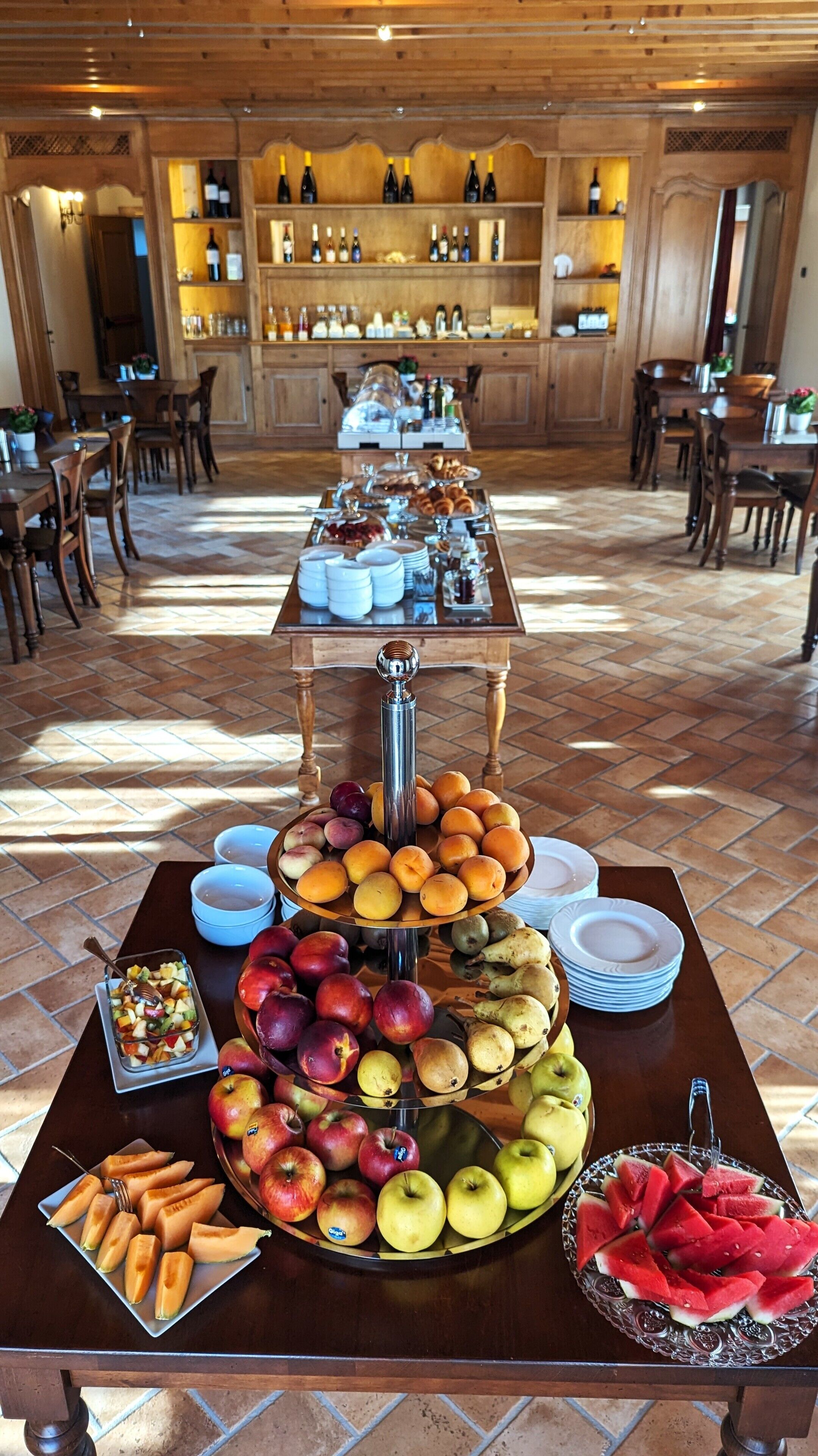 Una mesa de buffet con una variedad de frutas y pasteles, rodeada de muebles de madera y una zona de bar al fondo.