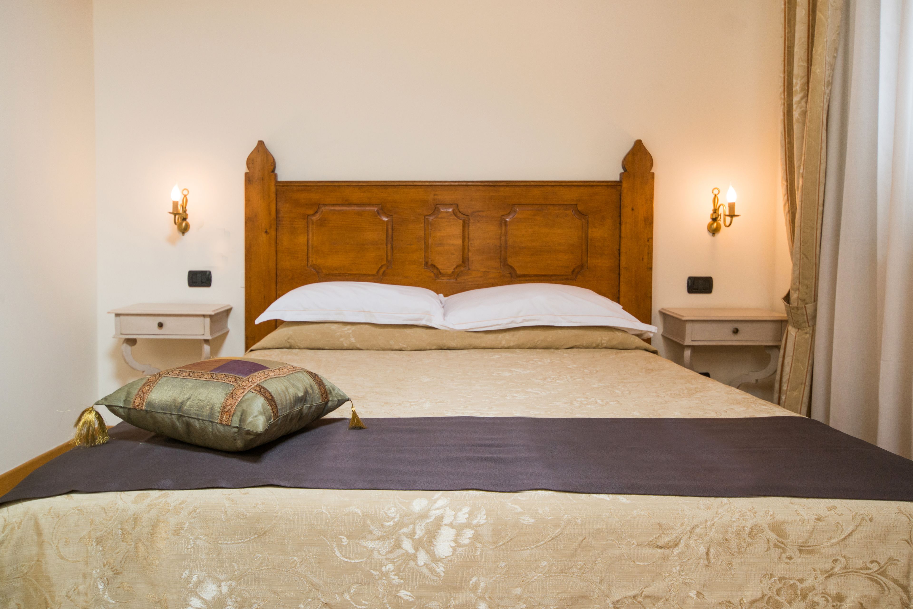 Una cama bien hecha con cabecera de madera, dos mesitas de noche con lámparas y un cojín decorativo.