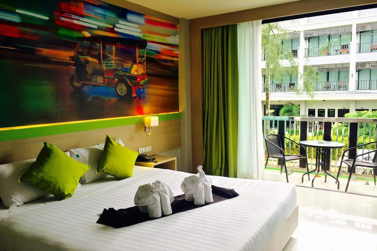 Habitación de hotel con una cama grande, almohadas verdes, un cuadro en la pared, un balcón con sillas y una mesa, y vista a un edificio.