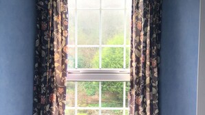 Rincón de una habitación con ventana, cortinas florales, una silla de madera con cojín y un cartel que dice "Thank you for not smoking". 