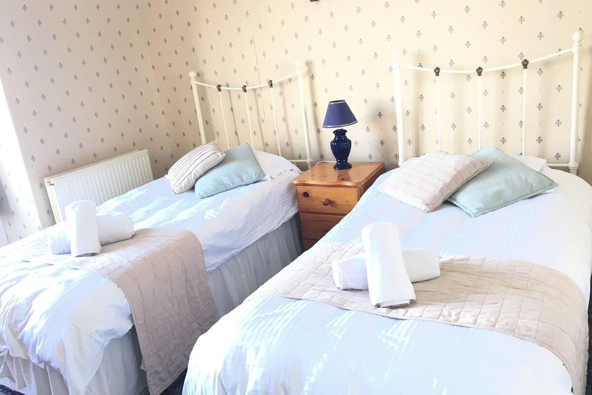 Une chambre avec deux lits, chacun équipé d’un oreiller et d’une serviette enroulée. Une table de chevet avec une lampe se trouve entre les deux lits.