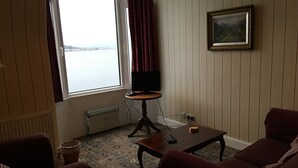 Una habitación con un ventanal grande, un televisor sobre un soporte de madera, una mesita pequeña y dos sofás rojos.