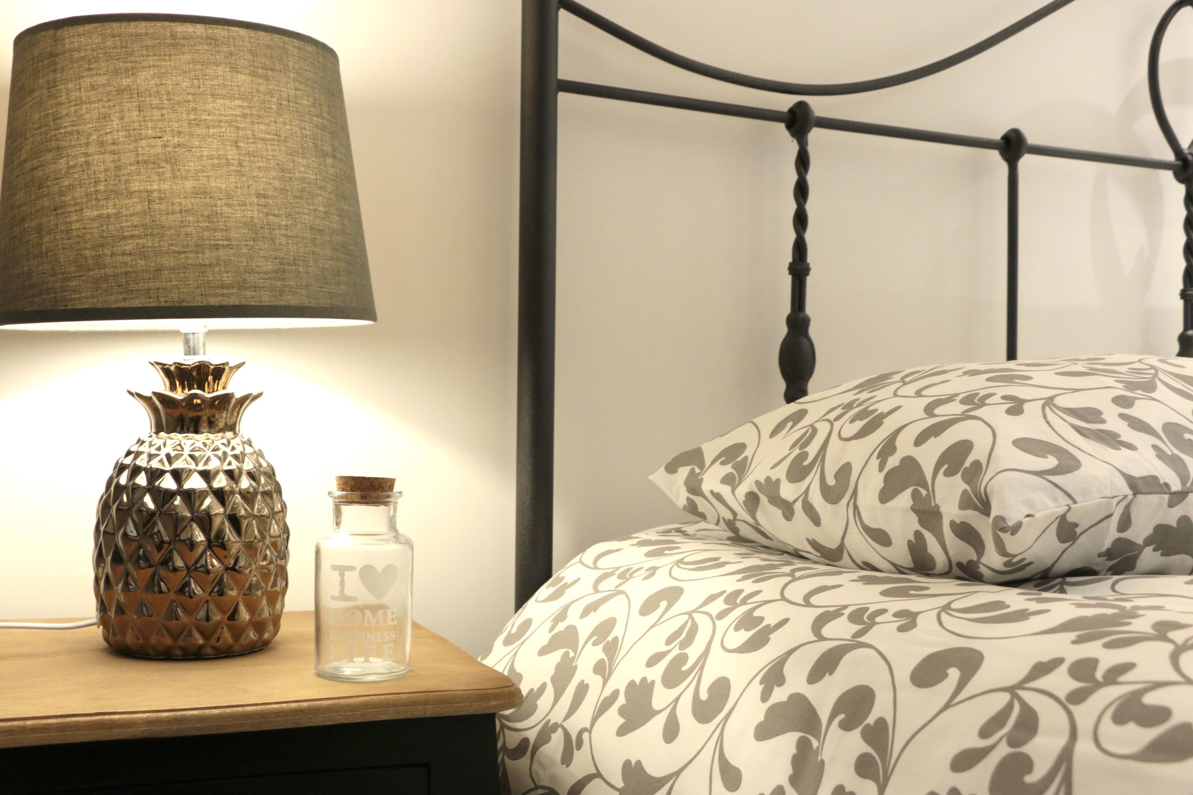 A bed with a patterned bedspread, a lamp with a textured lampshade, and a decorative vase on a bedside table.