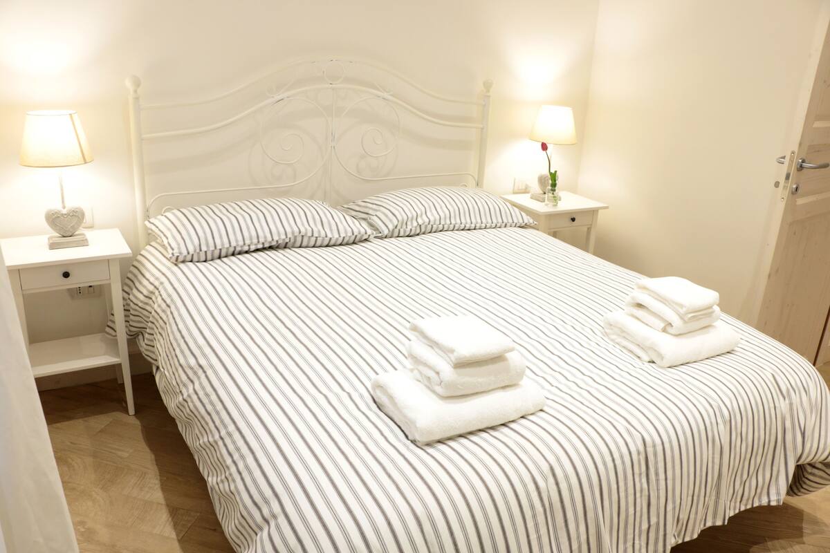 Una cama bien hecha con ropa de cama a rayas y dos toallas dobladas al pie. Hay dos mesitas de noche, una con lámpara y un adorno.