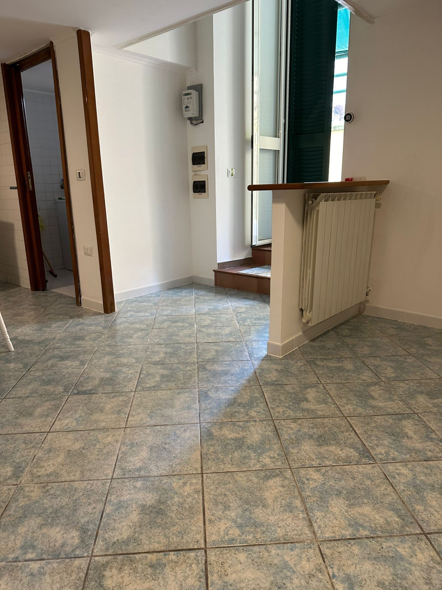 A spacious hallway with tiled flooring, a wooden door frame, a radiator, and a wall-mounted electrical box.