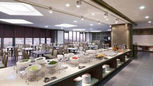 Un espace repas moderne, avec des tables et des chaises, un buffet proposant divers plats, et une vue sur la ville grâce à de grandes fenêtres.