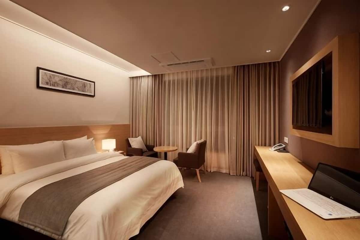 A hotel room with a large bed, a desk with a laptop, a chair, a small table, and a curtain.