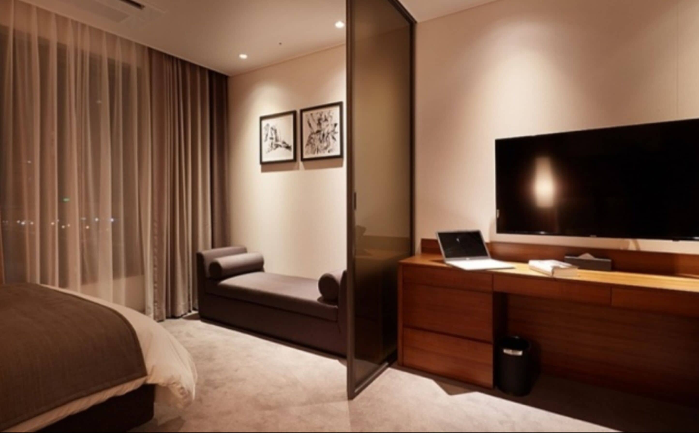 A modern hotel room with a bed, a desk with a laptop, a TV, and two framed pictures on the wall.