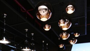 Un restaurante con una decoración moderna, que presenta una barra larga con sillas blancas tapizadas, una pantalla grande que muestra un paisaje montañoso y varias lámparas colgantes esféricas.