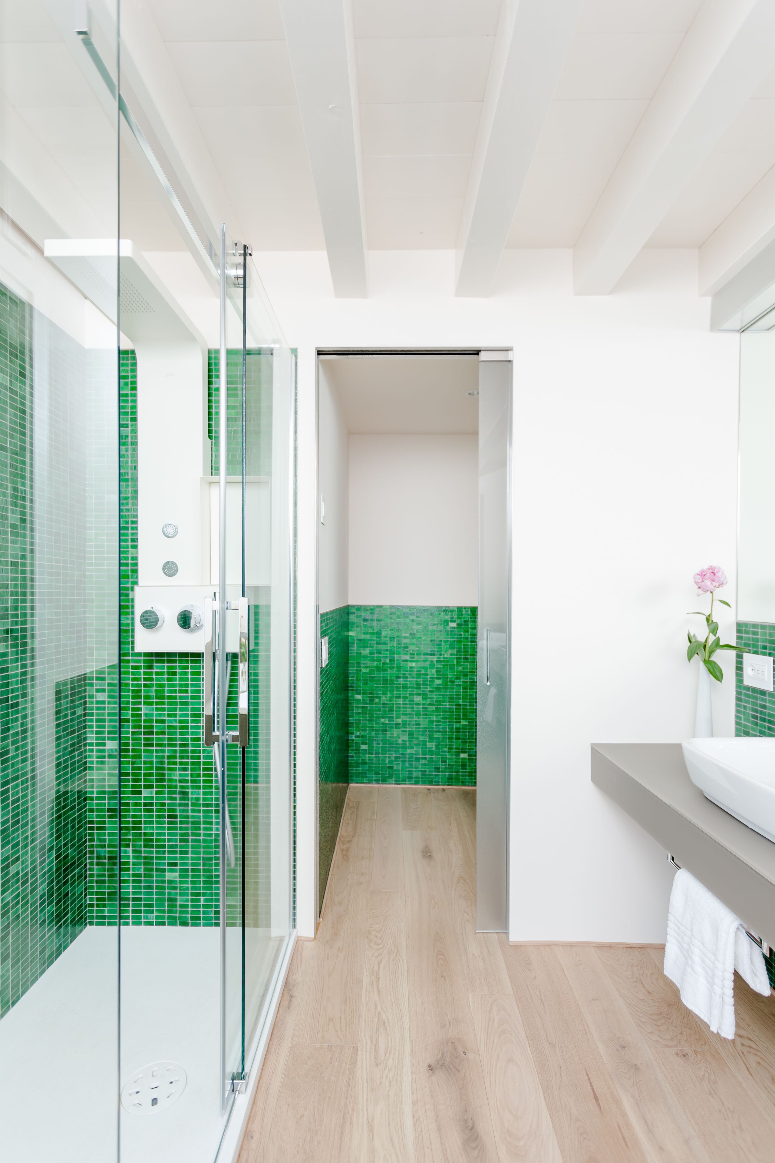 A modern bathroom with a glass shower enclosure, green mosaic tiles, a wooden floor, and a white sink with a vase of flowers.