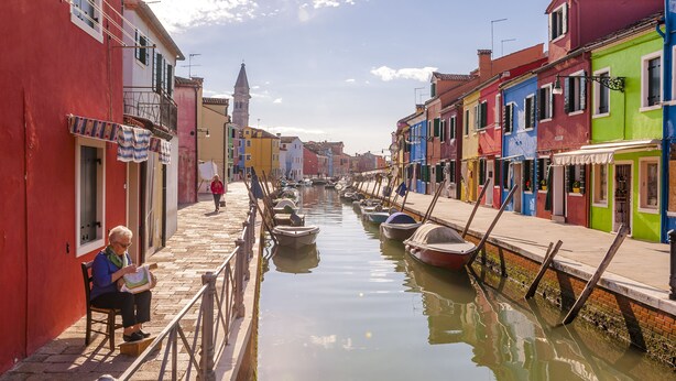 Un canal flanqueado por edificios coloridos, una mujer sentada junto al agua y una torre a lo lejos.