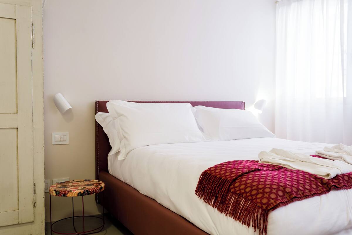 Una cama bien hecha con sábanas blancas y un plaid rosa estampado, una mesita redonda con un cubrecolorido, y una pared lisa con un interruptor de luz.