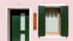 Un edificio rosa con puerta y contraventanas verdes, un buzón y un cartel con un número sobre la puerta.