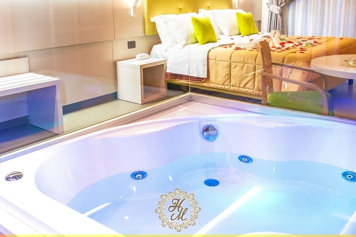 A modern hotel room with a large bed, a bedside table, a chair, and a hot tub with a logo.