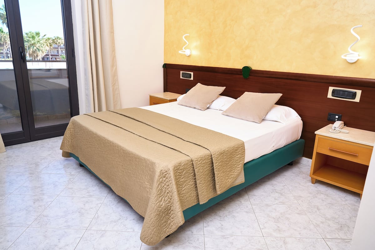 A hotel room with a bed, a nightstand, a bedside table, a window with curtains, and a view of the outside.