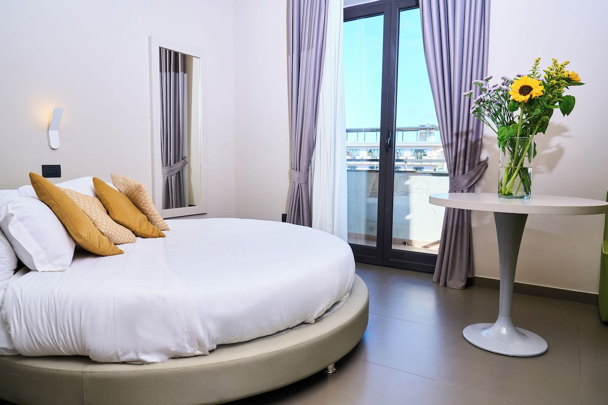 A hotel room with a large bed, a round table with a vase of flowers, and a view of a building through a window.
