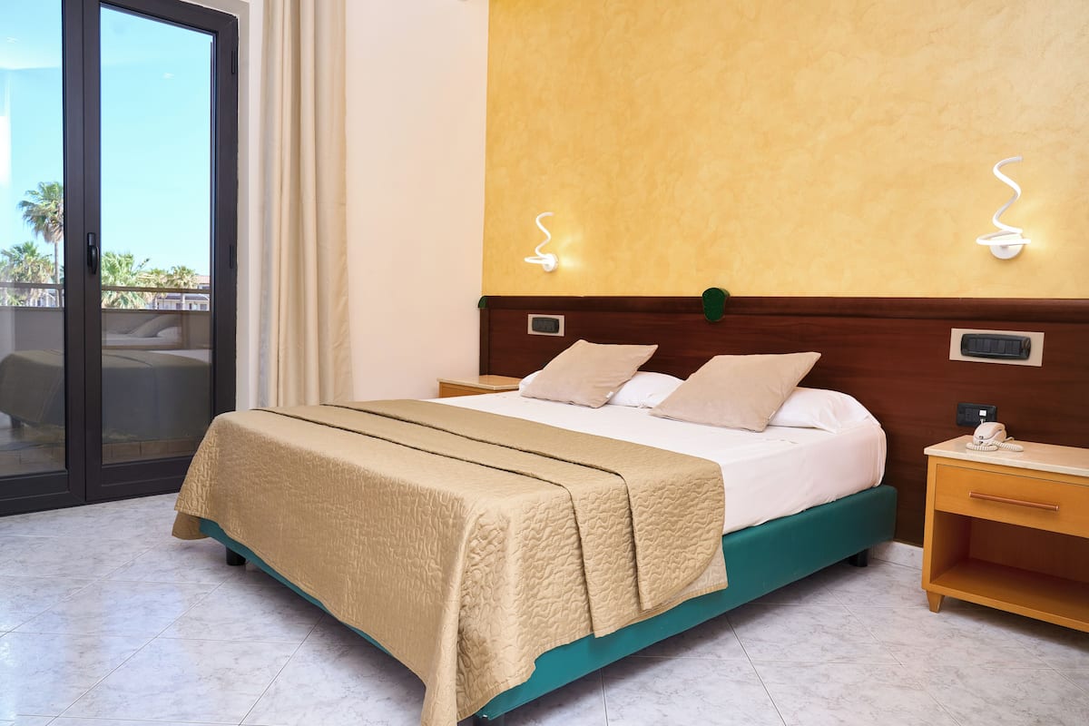 A hotel room with a bed, a nightstand, a wall-mounted light, and a sliding glass door leading to an outdoor area.