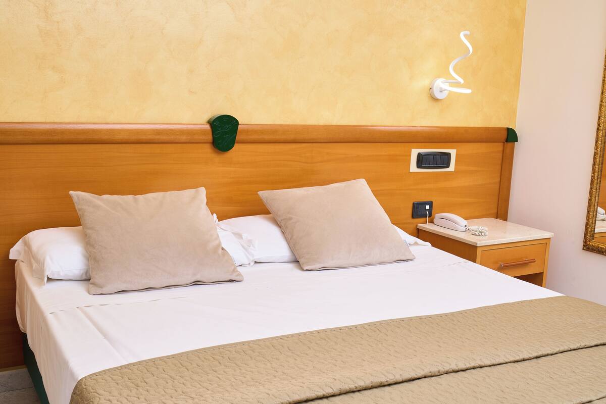 A hotel room with a bed, two pillows, a bedside table with a phone and a lamp, and a wall-mounted light fixture.