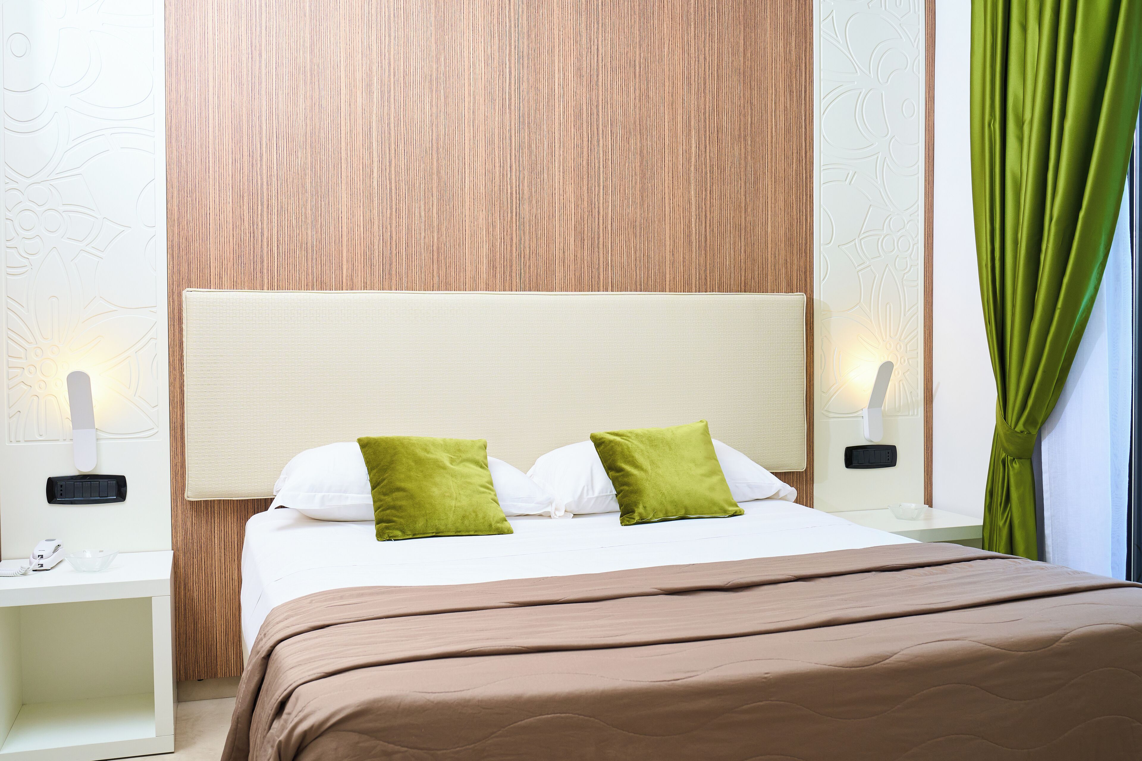 A neatly made bed with a brown bedspread and two green pillows, a wooden headboard, and a nightstand with a lamp and phone.