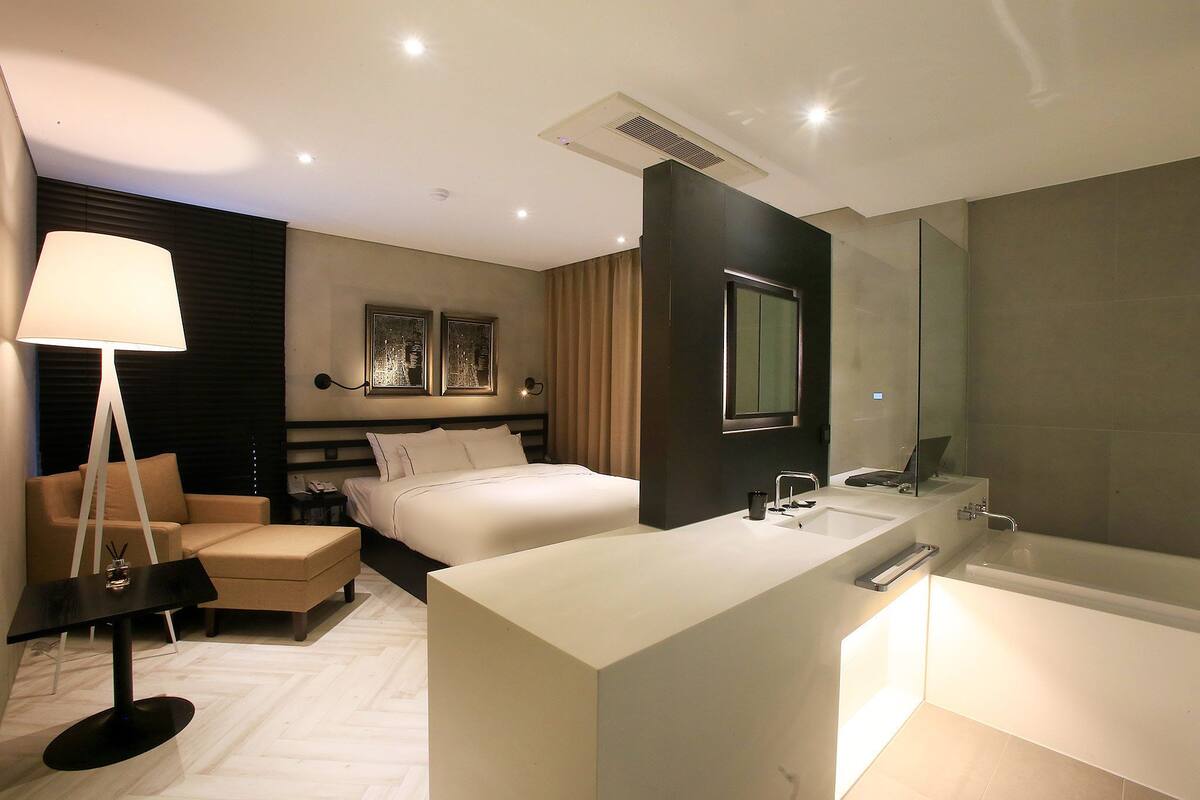 A modern hotel room with a large bed, a sitting area with a table and chair, a separate bathroom with a bathtub, and a large mirror.