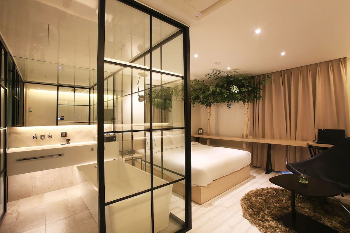 A modern bedroom with a glass partition, a bed, a desk, and a plant.